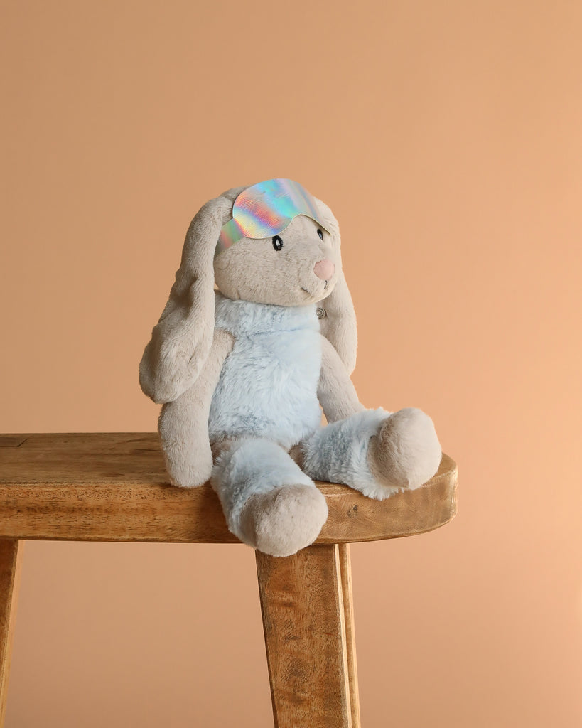 Steiff Hoppie Rabbit in Ski Outfit, 12 inches, is a light blue and gray plush bunny with floppy ears. Wearing a shiny iridescent sleep mask, this winter stuffed animal stands out against the plain light brown background.