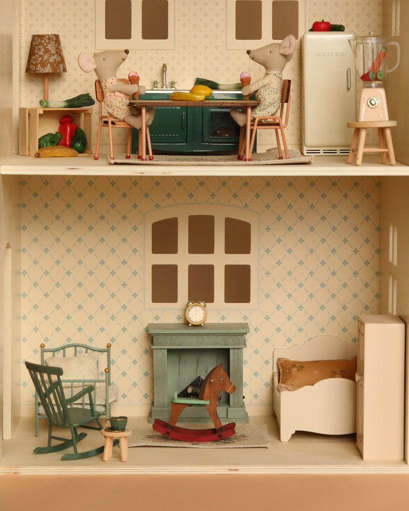 The Maileg Fully Furnished Holiday Dollhouse Set features two floors: upstairs, two toy mice sit at a kitchen table; downstairs are miniature furniture pieces including a green rocking chair, red rocking horse, crib, and fireplace with clock.