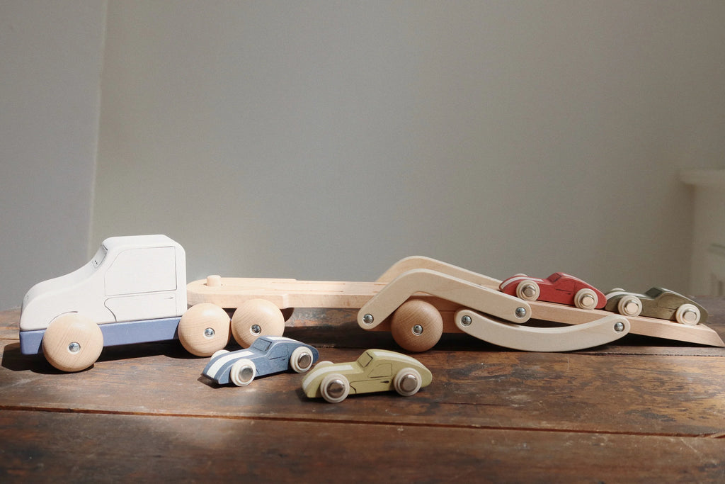 The Konges Sløjd Wooden Car Transporter is arranged on a wooden surface, its larger FSC-certified beech wood structure featuring an extending trailer that carries several colorful smaller cars. Sunlight illuminates the toys, creating a warm and playful atmosphere.