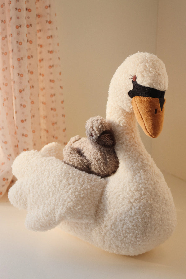 The Konges Sloejd Activity Music Swan features a plush swan with a brown bear nestled on its back, set against floral-patterned curtains, and plays Brahms' lullaby. Its soft, textured design creates a cozy ambiance, making it an ideal activity toy that provides a soothing melody.