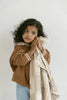 A young child with curly hair in a brown sweater and jeans rests their cheek on the Organic Cotton Baby Blanket - Pony, featuring crochet trim, while gazing thoughtfully to the side against a plain light background.