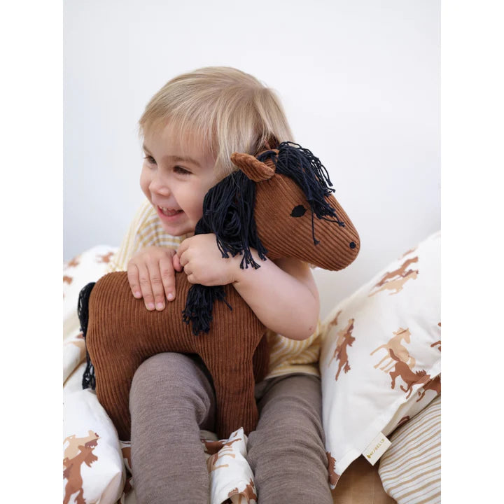 A toddler with light hair smiling joyfully while hugging a brown Fabelab Friends Stuffed Animal - Horse Hector toy, sitting against pillows with horse prints.