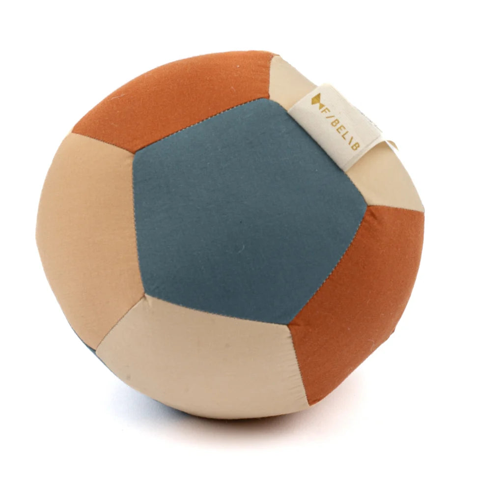 The Fabric Ball - Cobblestone is a soft, developmental baby toy made from pentagonal panels of muted blue, tan, cream, and rust organic cotton, with a fabric tag—ideal for sensory play and gentle exploration.