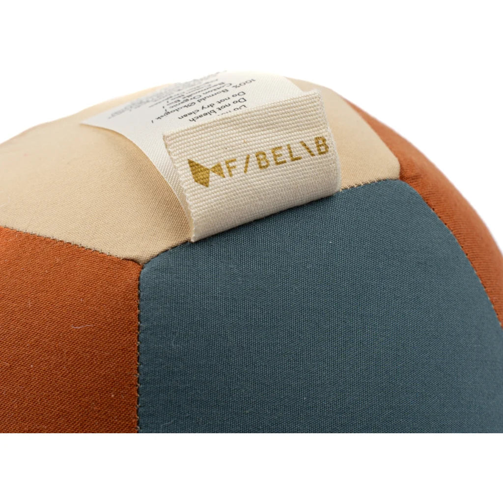 A close-up of the Fabric Ball - Cobblestone, an organic cotton baby toy with teal, rust, and beige panels. A white tag with gold "AF/ BELIB" text is sewn on top, making this a stylish developmental toy for babies.