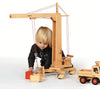 A young blonde-haired child is joyfully playing with a Fagus Wooden Crane, a handcrafted toy made in Germany. The wooden crane skillfully lifts a red block as a wooden toy truck sits nearby. The child beams with delight, fully engaged with the crane's controls.