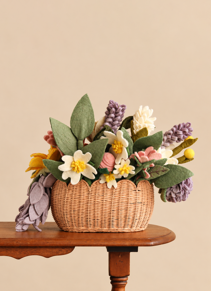 A small Basket of Handmade Felt Flowers - Final Sale filled with a wool felt bouquet of colorful flowers and green leaves, sitting on a wooden side table.