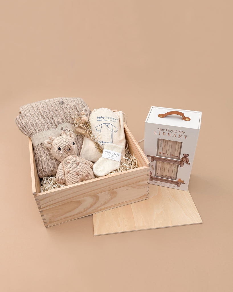 The Newborn Gift Set - Natural featuring a knitted baby blanket, Cuddle + Kind plush bear, socks, and bib sits beside an "Our Very Little Library" box on a beige background.