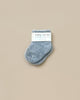 A pair of small, fuzzy gray baby socks with a "baby socks" label sits next to the whimsical Let's Go Explore Book from the Newborn Gift Set - Blue on a plain light beige background.
