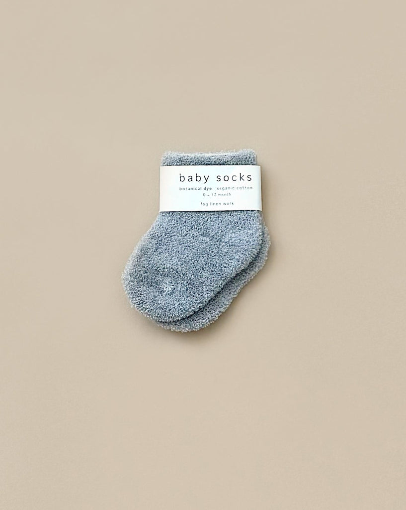 A pair of small, fuzzy gray baby socks with a "baby socks" label sits next to the whimsical Let's Go Explore Book from the Newborn Gift Set - Blue on a plain light beige background.