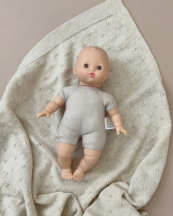 A Minikane Soft Body Doll (11") - Alice with a bald head, dressed in a gray onesie, lies on a textured light gray blanket with small eyelet holes. Made from phthalate-free vinyl, the doll has realistic features and a neutral expression, perfect for imaginative playtime.