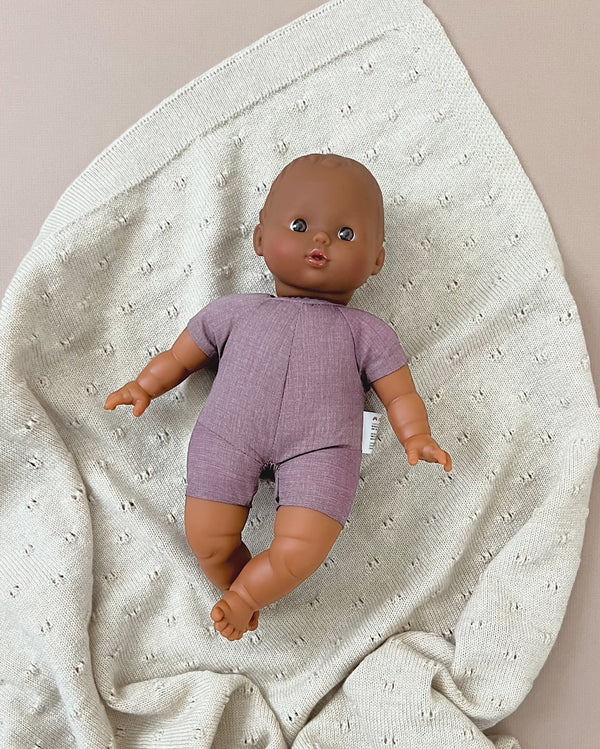 A Minikane Soft Body Doll (11") - Luke wearing a purple romper lays on a cream-colored, textured blanket spread out on a flat surface. The doll, made of phthalate-free vinyl, has a surprised expression, and its outfit contrasts with the soft, patterned fabric beneath it. Perfect as a bedtime companion.