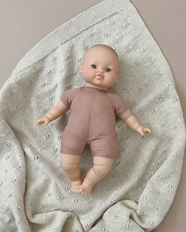 A Minikane Soft Body Doll (11") - Maé with blue eyes, dressed in a short-sleeve, pinkish-brown one-piece outfit, lies on a light grey, textured blanket. The phthalate-free vinyl doll rests on the soft, knitted fabric with small perforations, creating a cozy setting perfect for imaginative playtime.
