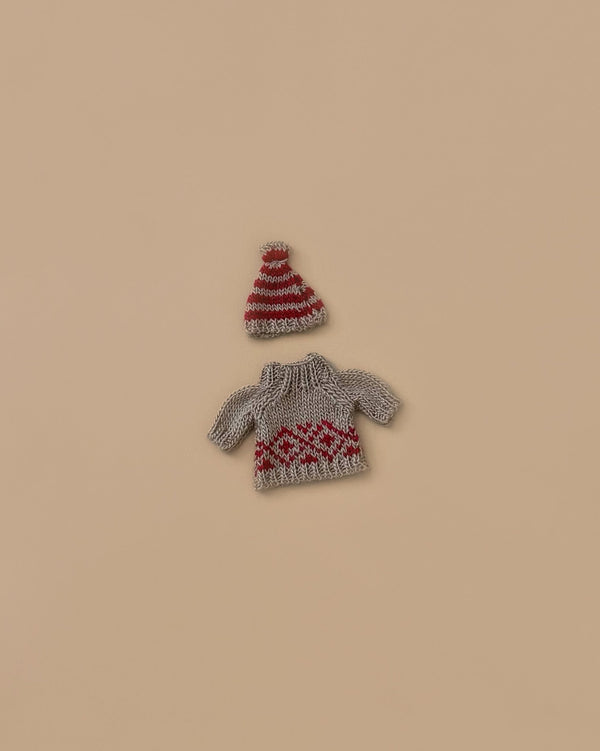 The Maileg Knitted Sweater and Hat for Mom Mouse features a tiny sweater with charming red patterns, complemented by a matching red and gray striped hat adorned with a pom-pom, making it an ideal winter wardrobe addition or an endearing accessory for your mouse. The set is presented against a simple beige background.