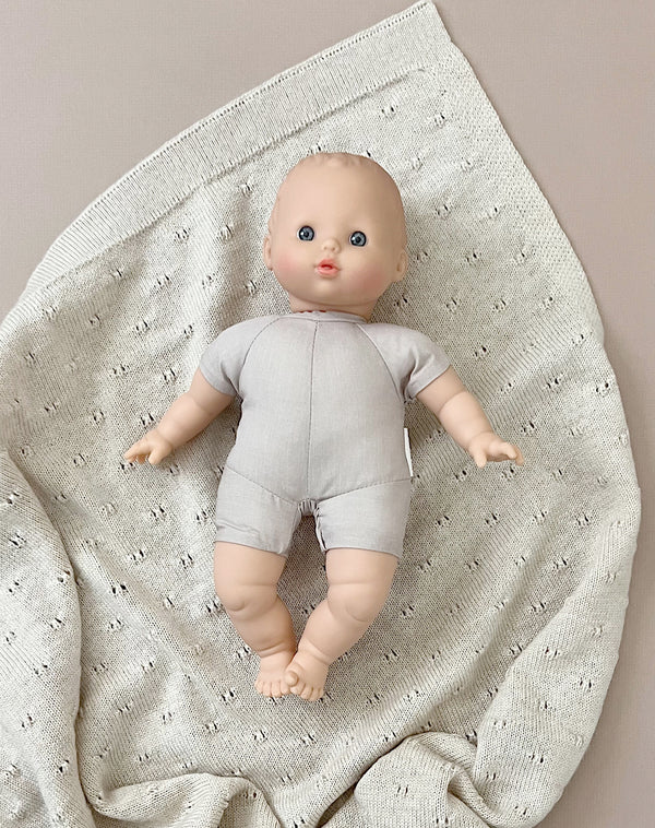 A Minikane Soft Body Doll (11") - Clarisse with blue eyes and light-colored hair is dressed in a light grey onesie. Made from phthalate-free vinyl, the doll lies on a cream-colored knitted blanket with small holes in a diamond pattern, perfect for imaginative playtime. The blanket is spread out on a beige surface.