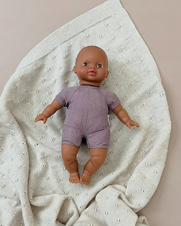 A Minikane Soft Body Doll (11") - Oscar is lying on a cream-colored knitted blanket. The bedtime companion wears a short-sleeved, lavender romper, and the phthalate-free vinyl doll rests on a blanket with small, evenly spaced holes in a pattern.