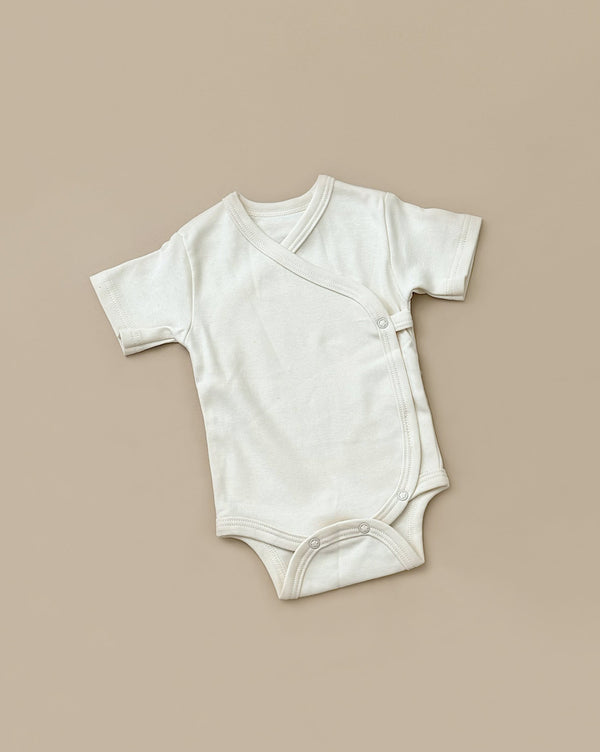 A Baby Romper with short sleeves lies flat on a beige background. This piece of baby clothing features a wrap-over design with snap buttons on one side for closure, crafted from soft, organic cotton.