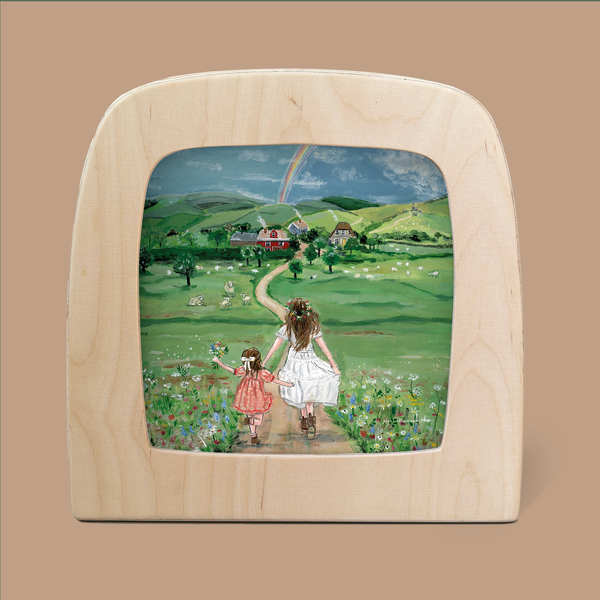 A silhouette from the "Silhouette Only - Rainbow Magic for Toverlux Lamp" set depicts a woman and child walking hand in hand through a lush green meadow. As they walk toward distant houses, a rainbow gracefully arches above them in the cloudy blue sky.