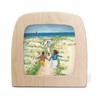 The "Silhouette Only - Sea" framed painting shows two children running hand in hand toward the beach, surrounded by sand dunes and grass under a bright blue sky. A lighthouse stands in the distance against the ocean backdrop. The frame is crafted from light wood with food-safe materials.