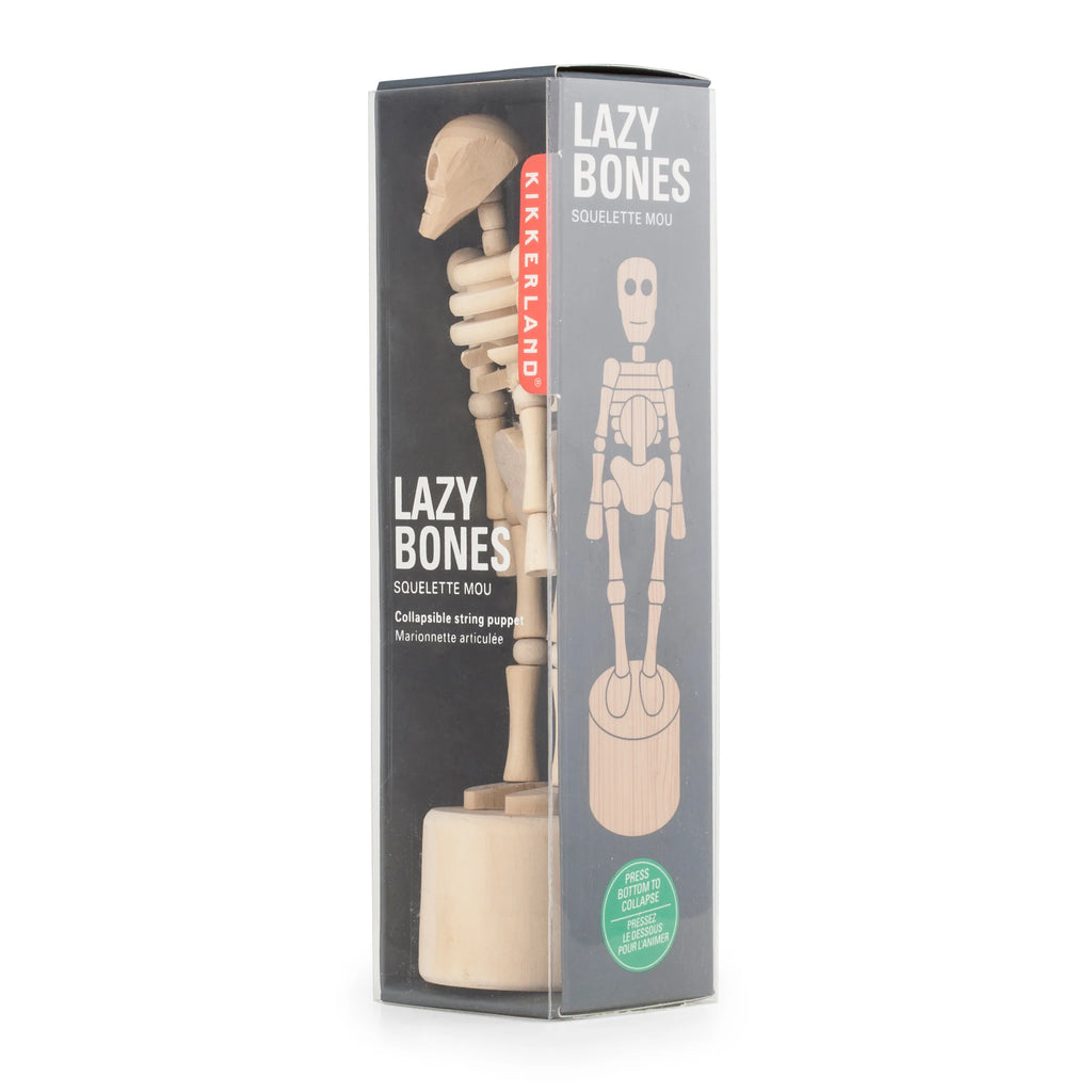 The image displays a packaged "Wooden Lazy Bones Skeleton Pop Up" by Kikkerland. The box features an illustration of the standing skeleton puppet on the right side and a clear window on the left, revealing the actual wooden sculpture inside.