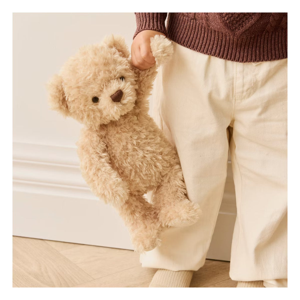 A young child in cream pants and a brown sweater holds Snuggle Bunnies - Arthur the Bear (ships in approximately one week), standing on a wooden floor by a white wall.