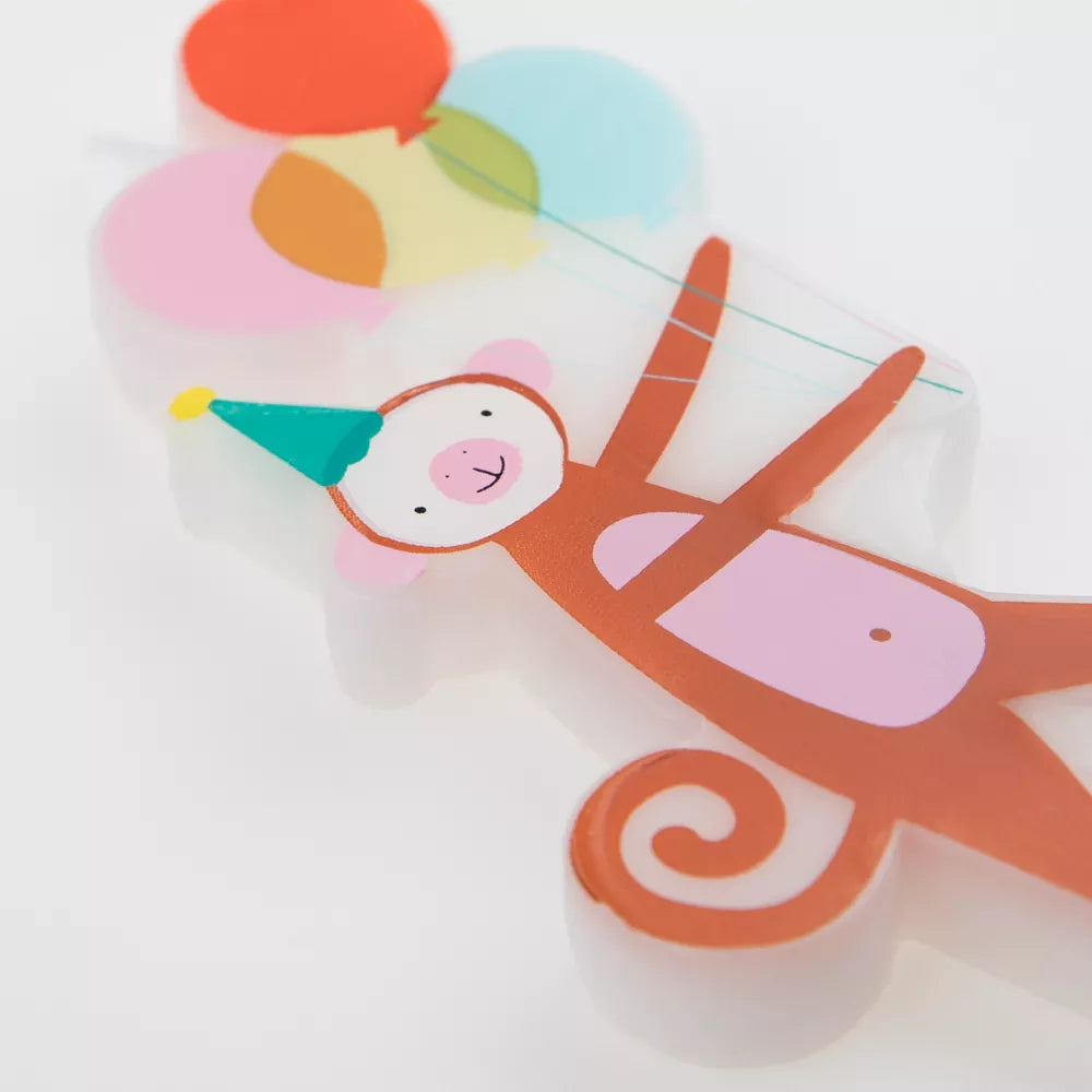 A set of Meri Meri Animal Parade Candles shaped like a smiling monkey wearing a party hat, designed for a baby shower, set against a translucent backdrop with vibrant, overlapping geometric shapes.