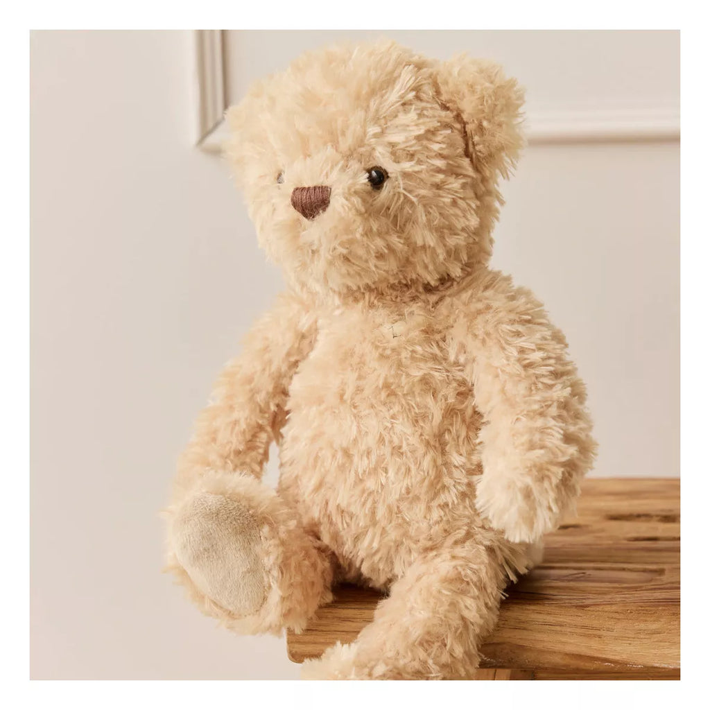 Arthur the Bear from Snuggle Bunnies, made of plush fabric, sits on a wooden surface against a light wall. This soft, light brown bear with a dark brown nose is perfect for cozy hugs and ships in about one week.