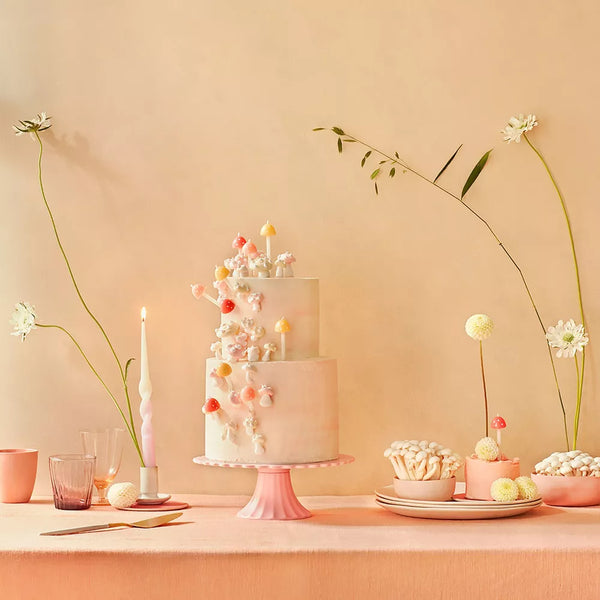Elegant dessert table setup for a fairy garden party with a three-tiered cake adorned with delicate pastel flowers, surrounded by Meri Meri Mushroom Birthday Candles, flowers in vases, and bowls of sweets on a peach.