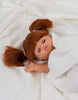 A small Minikane Baby Doll With Sleeping Eyes (13") - Raphaëlle with brown pigtails lies wrapped in a white blanket. The doll has a soft body, closed eyes, and a peaceful expression on its face, emitting a natural vanilla scent.