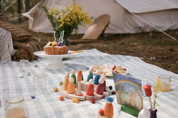 A whimsical outdoor birthday party setup with a birthday cake on a stand, various colorful Grapat Your Day figures, toys, and a canvas tent in the background, all laid out on
