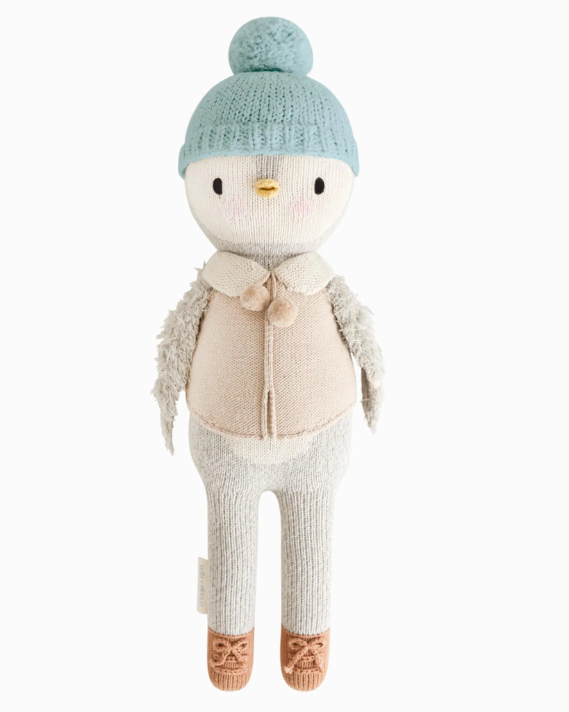 Cuddle + Kind Greyson the Penguin is a fair trade plush toy featuring a light blue knit hat with pom-pom, beige vest with pom-pom buttons, brown shoes, gray wings, yellow beak, and black stitched eyes.