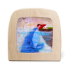 The felt art piece, "Silhouette Only - Whale Friend for Toverlux Lamp," depicts a child in a red sweater lying on grass and reaching toward a blue whale surfacing from the water, with birds flying above. This enchanting scene is framed in a wooden, TV-shaped frame to charm any home atmosphere.