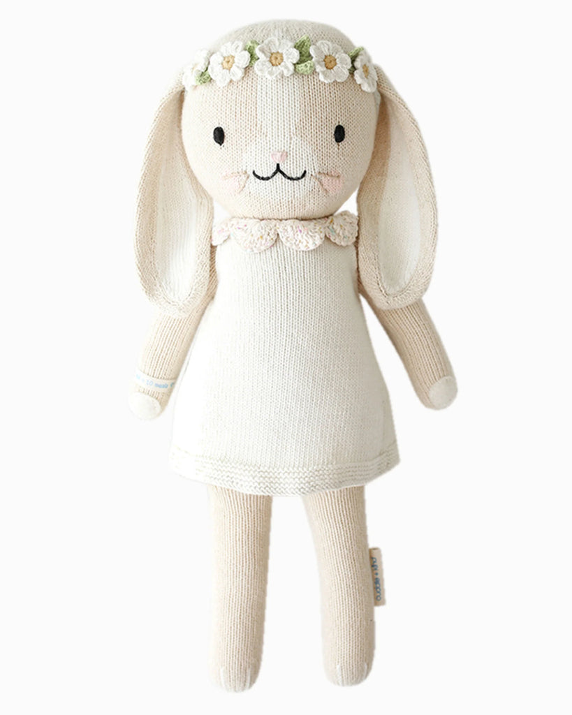 Cuddle + Kind Hannah the Bunny is a cream-colored hand-knit bunny doll with long floppy ears, dressed in a white dress and daisy flower crown. Made from premium cotton yarn, this fair trade bunny has black eyes, pink cheeks, and a sweet smile.