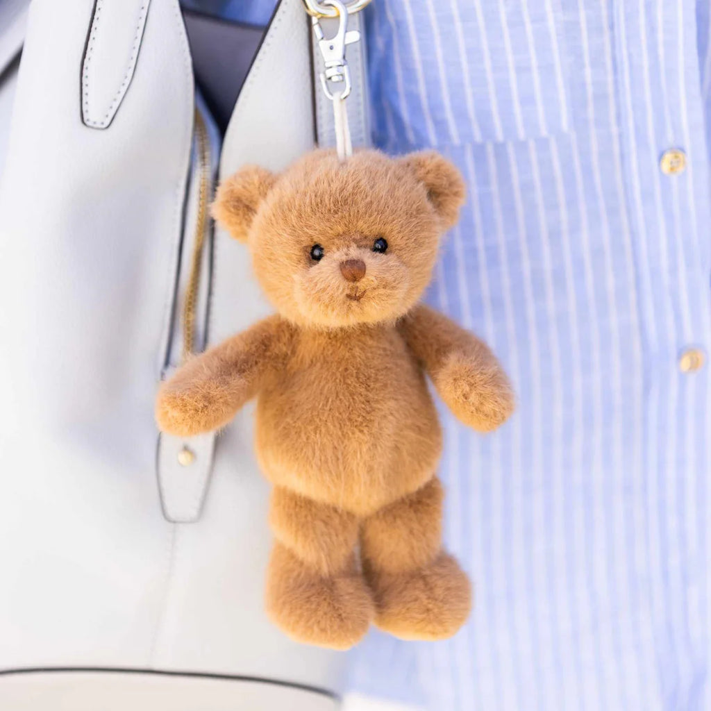 The Bukowski - Happy Mavi Bag Charm XL, a small fluffy bear plush keychain, is attached to a white handbag, with someone in a light blue striped shirt in the background.