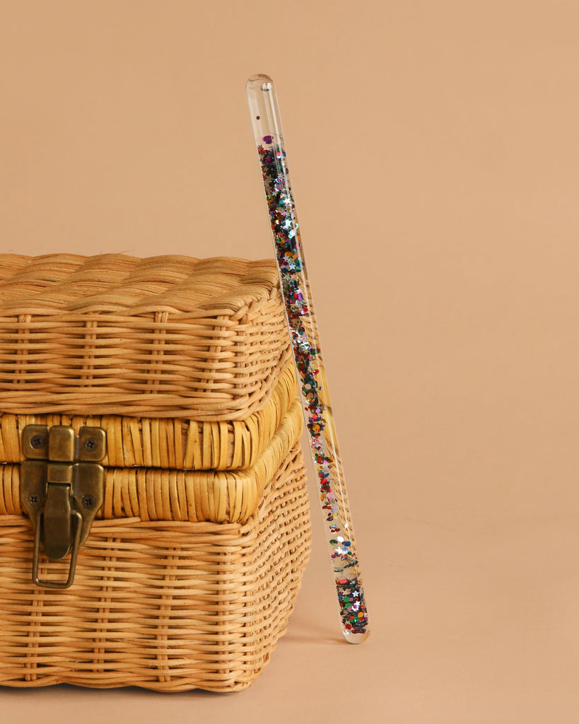 The Konges Sloejd Magical Glitter Wand - Star Glitter, filled with colorful glitter, rests against a woven wicker basket with a brass latch on a plain beige background.