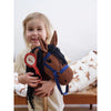 A young child with blonde hair smiles while holding a Hobby Horse with a blue bridle and black mane. The horse has a red ribbon attached to it. In the background, there is a bed with horse-patterned bedding made from soft organic cotton.