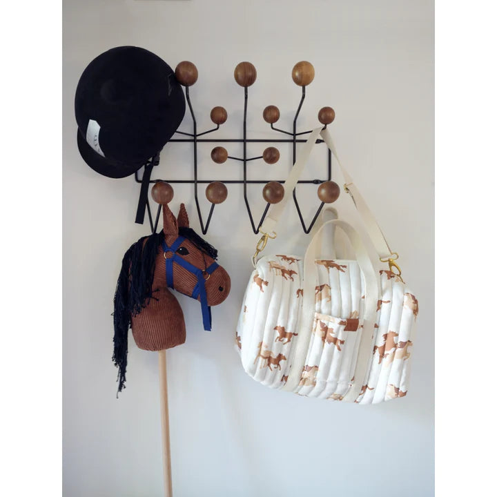 A black riding hat is hung on a wall-mounted coat rack with wooden knobs. Next to it, there's a brown Hobby Horse with a blue bridle and black mane. A white quilted bag made of organic cotton by Fabelab, adorned with a horse print, hangs on the same rack.