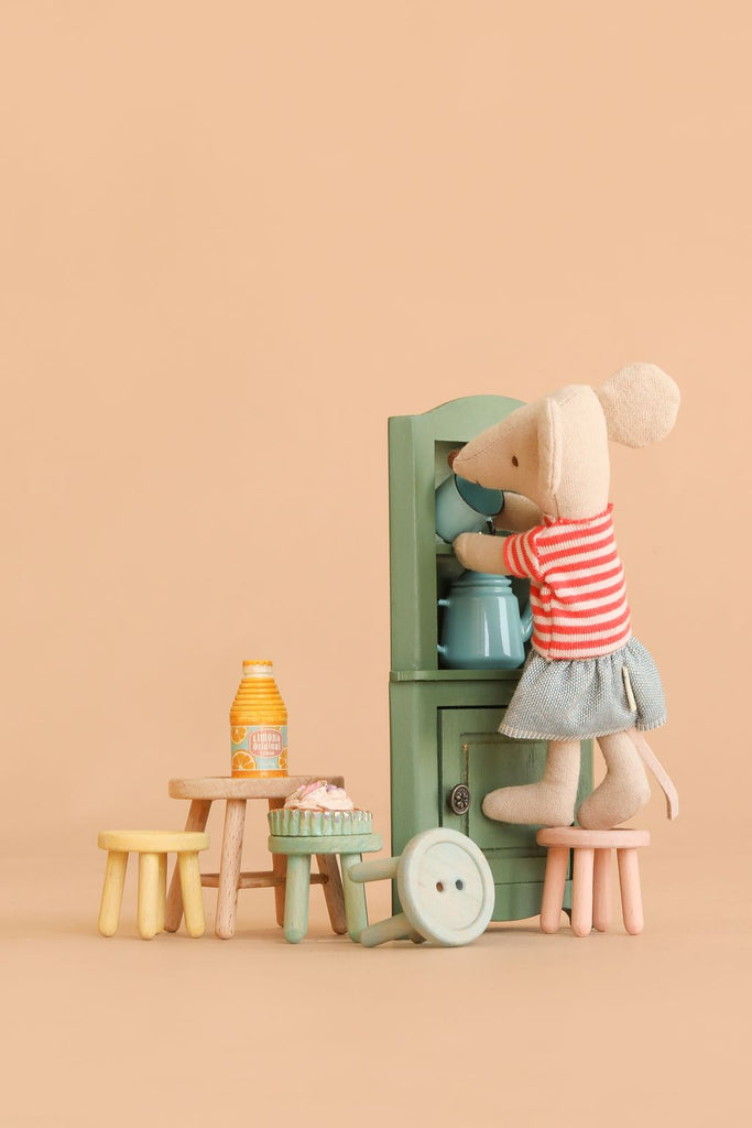 A toy mouse in a striped shirt and skirt stands on the Maileg Stool Set, Mouse - Light Rose/Light Mint, reaching into a green cabinet with a charming vintage paint finish. Small stools from the set, a bottle, and a plate with pastries are nearby, all delicately arranged on a beige background.