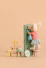 A toy mouse in a red-striped shirt and blue skirt stands on the Maileg Stool Set, Mouse – Mint/Light Yellow, reaching into a green cabinet with a charming vintage paint finish. Miniature tables hold a bottle, cupcakes, and a large button on a beige background.