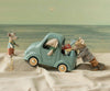 Three toy mice, dressed in clothes, engage in imaginative play with the PRE-ORDER: Maileg Truck, Mouse (Estimated Arrival Early November) on a sandy surface. One mouse drives the mouse family truck, another pushes from behind, and the third stands nearby. The playful scene is set against a painted backdrop of sea and sky.
