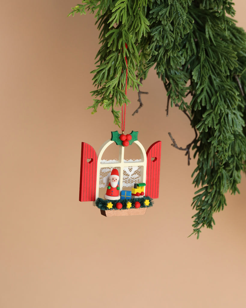 Hanging from a green evergreen branch is the Christian Ulbricht Ornament - Window With Santa, featuring open red shutters that reveal a miniature Santa, a wreath, and colorful presents. Crafted in Germany, it is set against a warm beige background.