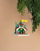 The Christian Ulbricht Ornament - Nativity, crafted in Germany, is shaped like a festive house adorned with snow-covered green trees. Hanging from a branch against a beige background, it includes small figures and a yellow flag on top, perfectly capturing the essence of holiday charm.