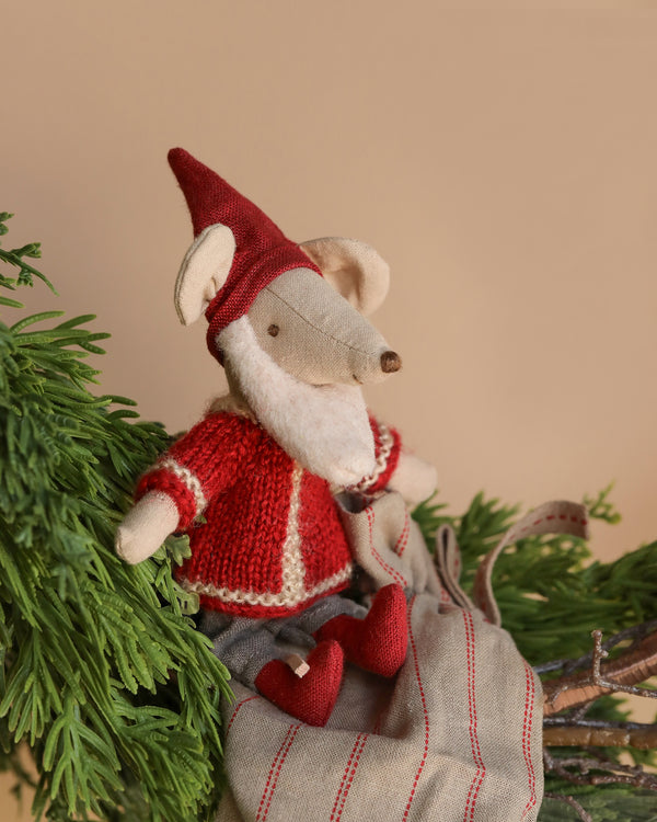 The Maileg Santa Mouse, a small plush mouse dressed in festive attire, sits on a green branch beneath the twinkling Christmas night. This charming big brother holds a striped sack and adds holiday cheer to the scene with his pointy hat and joyful spirit.