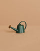 A Maileg Woodland Picnic Set, featuring a small dark green watering can with a curved handle and long spout, sits beside charming wooden mushrooms on a plain beige surface against a matching beige background.