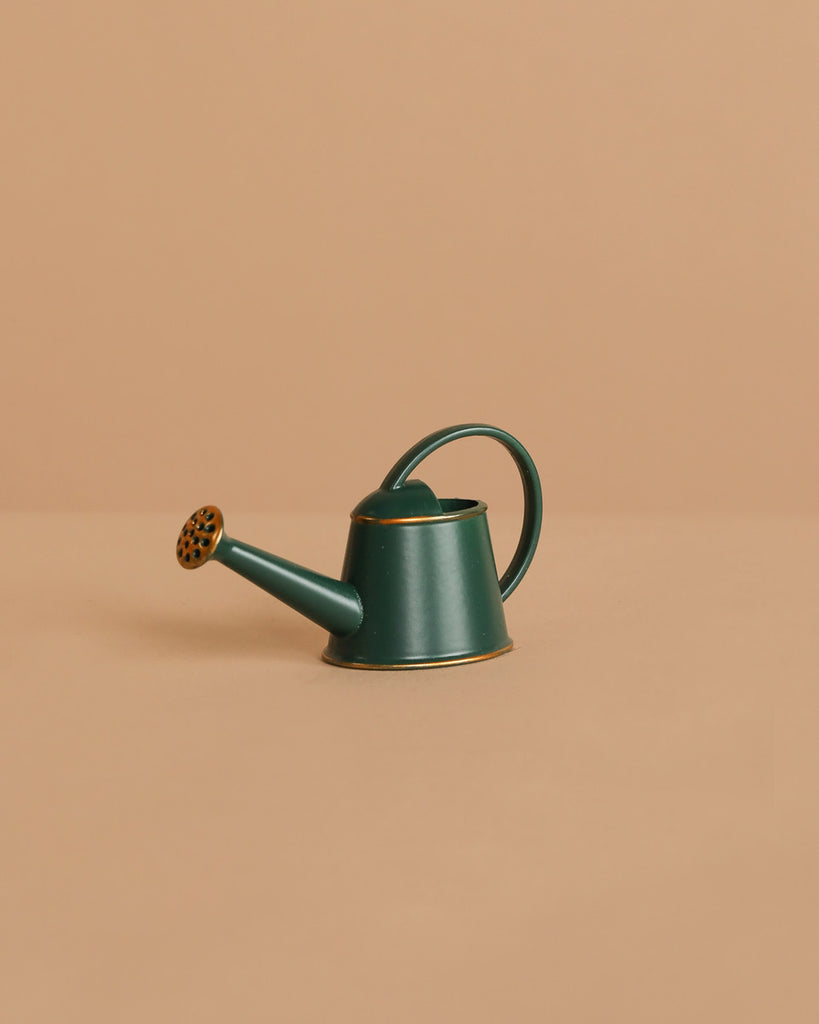 A Maileg Woodland Picnic Set, featuring a small dark green watering can with a curved handle and long spout, sits beside charming wooden mushrooms on a plain beige surface against a matching beige background.