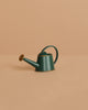 A small, green metal watering can with a round handle and spout sits on a beige surface, perfect next to the Maileg Living Room Set, against a matching beige background.