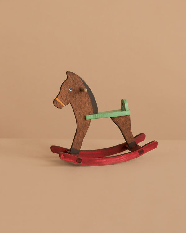 The Maileg Mouse Rocking Horse, featuring a brown body, green handle, and red rockers, rests on a beige surface—ideal for tiny mice or magnetic-handed figures to play and enjoy.