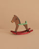 The Maileg Mouse Rocking Horse, featuring a brown body, green handle, and red rockers, rests on a beige surface—ideal for tiny mice or magnetic-handed figures to play and enjoy.