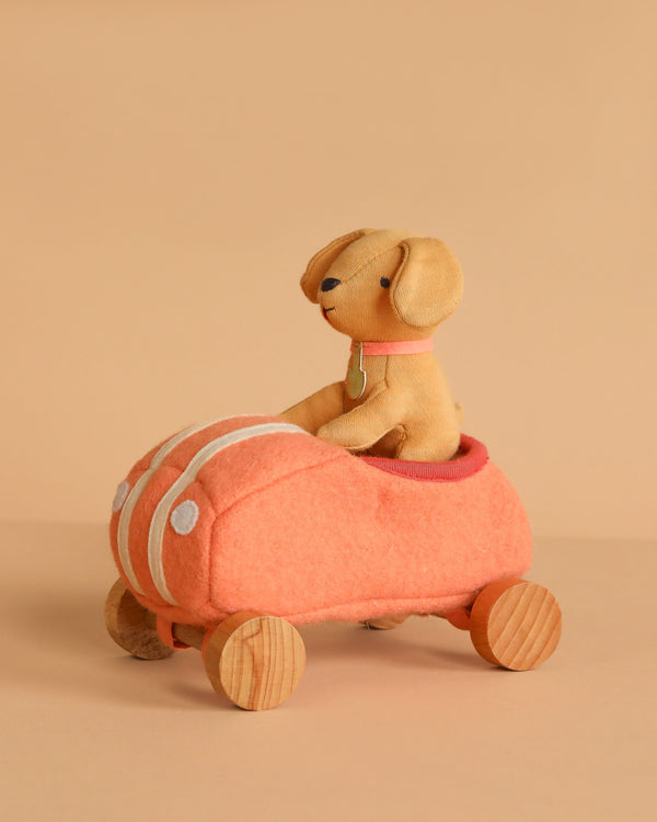 The Olli Ella Holdie Dog-Go - Racer Girl - Pink features a stuffed toy dog with a collar in a pink car with wooden wheels, sparking imaginative play on a beige background.