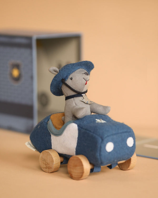 The Olli Ella Holdie Dog-Go Officer, a small plush toy, sits in a blue fabric police car with wooden wheels—perfect for imaginative play. The set is displayed on a beige background with its box behind it.