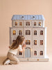 A young child kneels and peers inside the Fully Furnished Dollhouse With Lights, House of Monaco — The Grand Mansion, its illuminated windows glowing against a plain beige background.
